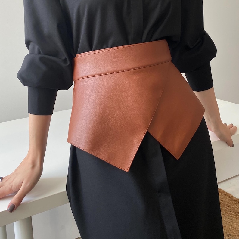 May include: A wide, tan-colored leather belt with an asymmetrical design. The belt is worn over a black dress, creating a layered look. The belt's texture appears smooth, and the color is a warm, earthy tone.