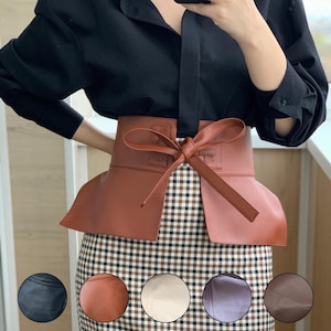 May include: A brown leather belt with a bow detail. The belt is worn over a plaid skirt and a black blouse. The belt has a wide design and a bow closure. The image also shows color swatches.
