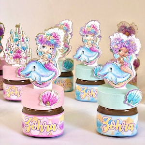 Peut inclure: Un ensemble de petits pots en verre remplis de tartinade au chocolat, décorés de surmatelas sur le thème des sirènes. Chaque pot a un couvercle rose, bleu ou vert et une étiquette avec le nom "Zohra" en lettres jaunes.
