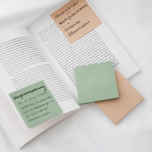 Può includere: Un libro aperto con diversi post-it colorati, tra cui verde, beige e pesca. Le note contengono testo in tedesco. Le pagine del libro sono bianche. Tema: studio.