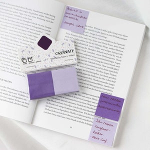 Könnte beinhalten: Eine Sammlung violetter Haftnotizen, einige davon auf den Seiten eines offenen Buches. Die Notizen sind mit Text beschriftet, und eine Packung der Haftnotizen ist ebenfalls sichtbar. Die Buchseiten sind weiß.