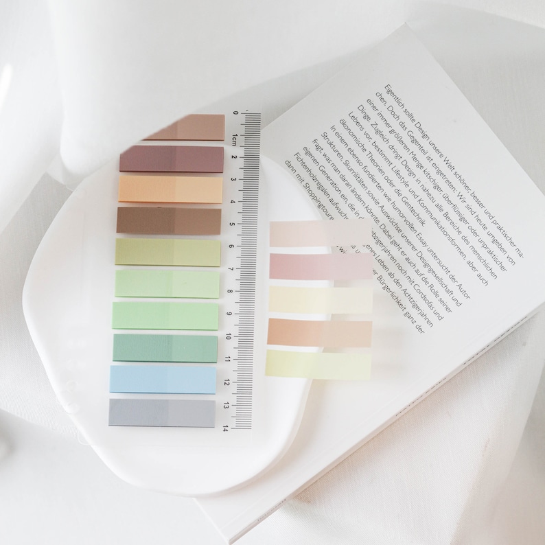 May include: A collection of rectangular sticky notes in various pastel colors, including shades of brown, green, and blue. A clear ruler with centimeter markings is placed next to the sticky notes. The notes are arranged on a white surface.