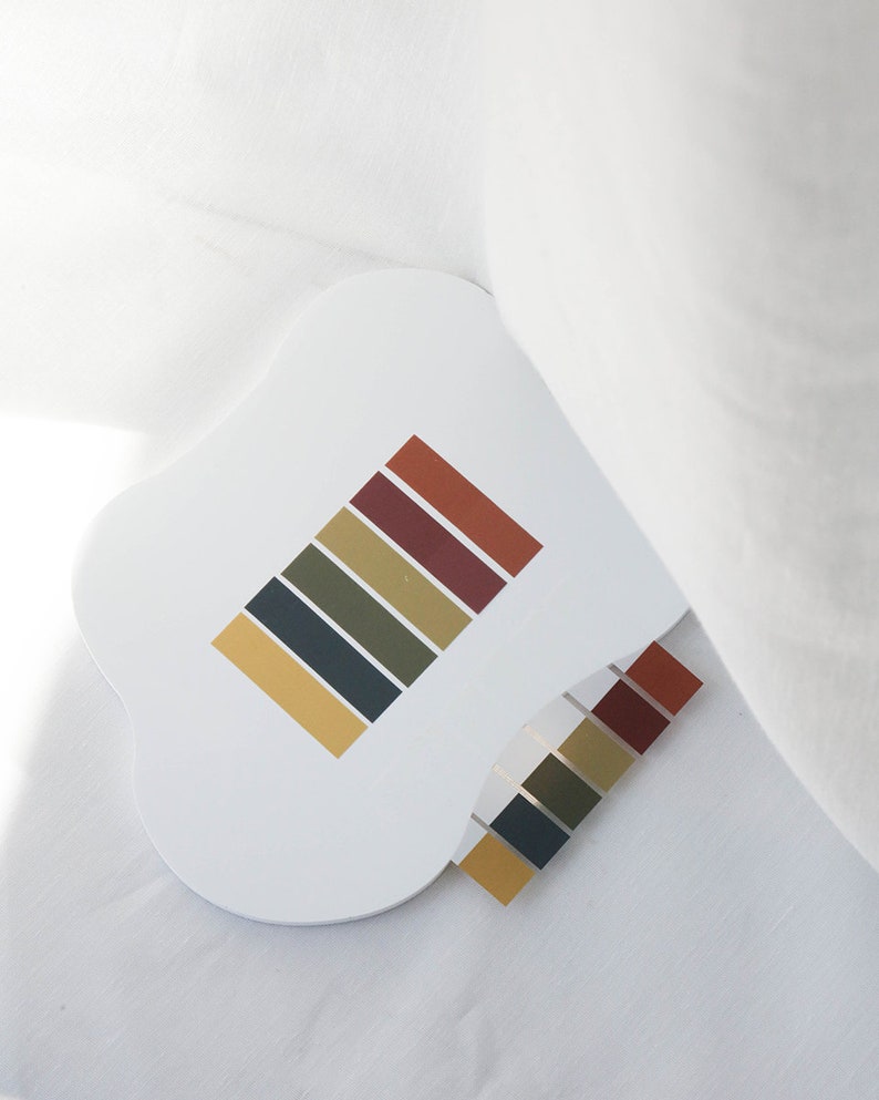 May include: A white, uniquely shaped coaster with a color palette design. The design features rectangular blocks in shades of gold, olive green, dark blue, and burgundy. A smaller, matching color palette is also visible.
