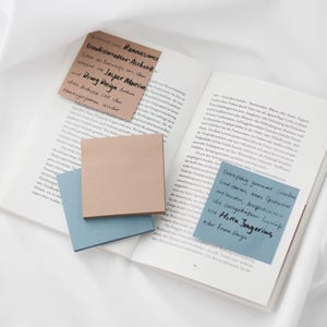 Könnte beinhalten: Ein offenes Buch mit mehreren Haftnotizen in verschiedenen Farben. Die Notizen haben handschriftlichen Text in schwarzer Tinte. Eine Notiz ist braun, zwei sind blau und eine ist hellpfirsichfarben. Die Seiten des Buches sind weiß.