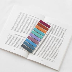 Puede incluir: Un libro abierto con notas adhesivas rectangulares de colores que marcan las páginas. Las notas adhesivas son de tonos morados, azules, verdes, naranjas y rojos. El libro está abierto en una página con texto.