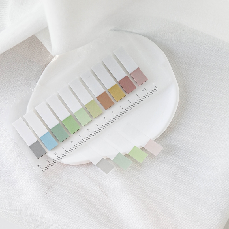 May include: A set of rectangular sticky notes with a white top and a colored bottom in shades of gray, blue, green, brown, yellow, and pink. A clear ruler with centimeter markings is placed across the notes on a white plate.