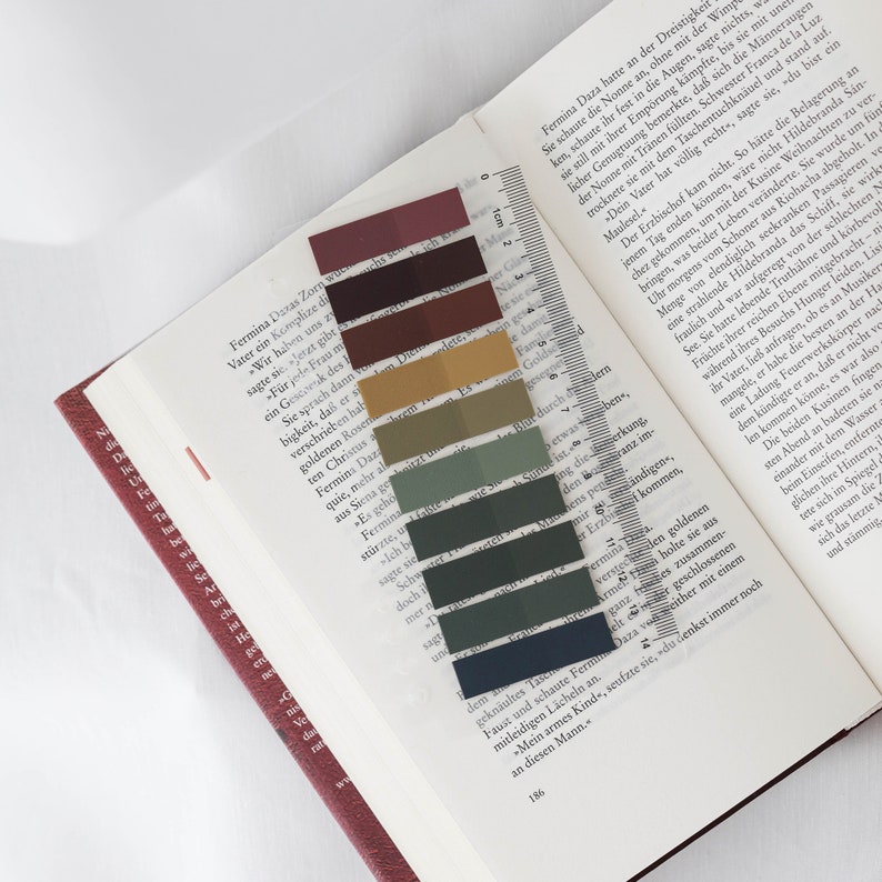 May include: A book with a clear ruler and colorful sticky notes. The sticky notes are in shades of burgundy, brown, olive green, and navy blue. The book is open to a page with German text.