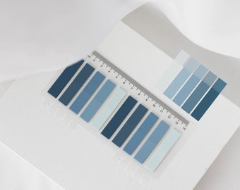 May include: A clear ruler with a blue and white color gradient sticky note set. The ruler is marked in centimeters and inches. The sticky notes are arranged in a row, with the darkest blue at the top and the lightest blue at the bottom.