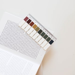 May include: A set of colorful sticky page markers with a clear plastic ruler, resting on the corner of an open book. The markers are rectangular, in shades of burgundy, brown, and green. The ruler is marked in inches and centimeters.