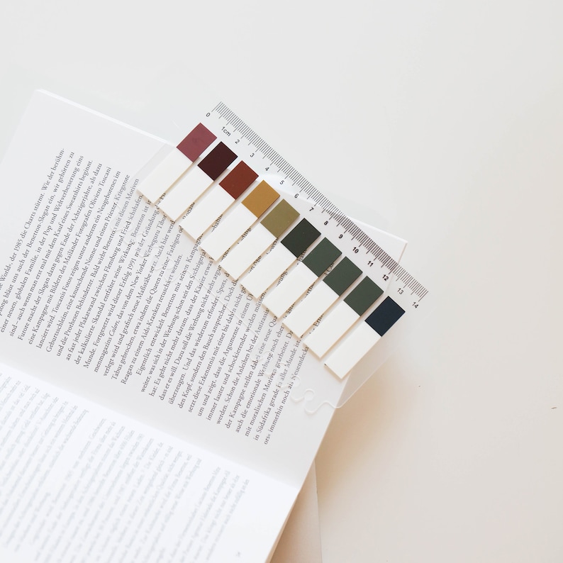 May include: A set of colorful sticky page markers with a clear plastic ruler, resting on the corner of an open book. The markers are rectangular, in shades of burgundy, brown, and green. The ruler is marked in inches and centimeters.