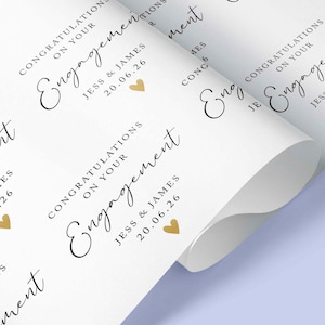 Op de afbeelding: Wit cadeaupapier met een herhaald patroon van het woord "Engagement" in elegant schrift, samen met de namen "Jess & James" en de datum "20.06.26", en kleine gouden hartaccenten. Het papier is opgerold.