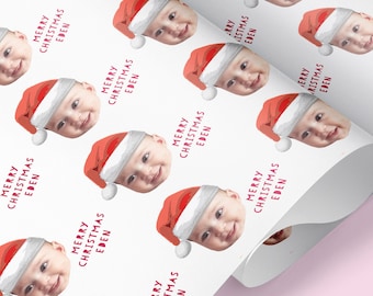 May include: White wrapping paper with a repeating pattern of a baby's face wearing a red Santa hat. The text "Merry Christmas Eden" is repeated throughout the pattern.