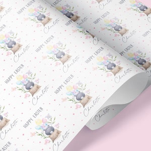 May include: White wrapping paper with a repeating pattern of a gray bunny holding pastel colored Easter eggs, pink flowers, and green leaves. The text "Charlotte" and "Happy Easter" is repeated throughout the pattern.