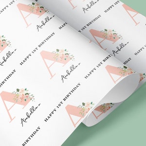 May include: White wrapping paper with a repeating pattern of a pink letter A with floral accents and the text "Arabella... Happy 1st Birthday".