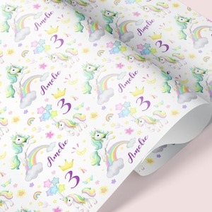 Op de afbeelding: Wit inpakpapier met een herhalend patroon van kleurrijke eenhoorns, regenbogen, sterren en bloemen. De naam "Amelie" en het nummer "3" worden herhaald in het hele patroon.