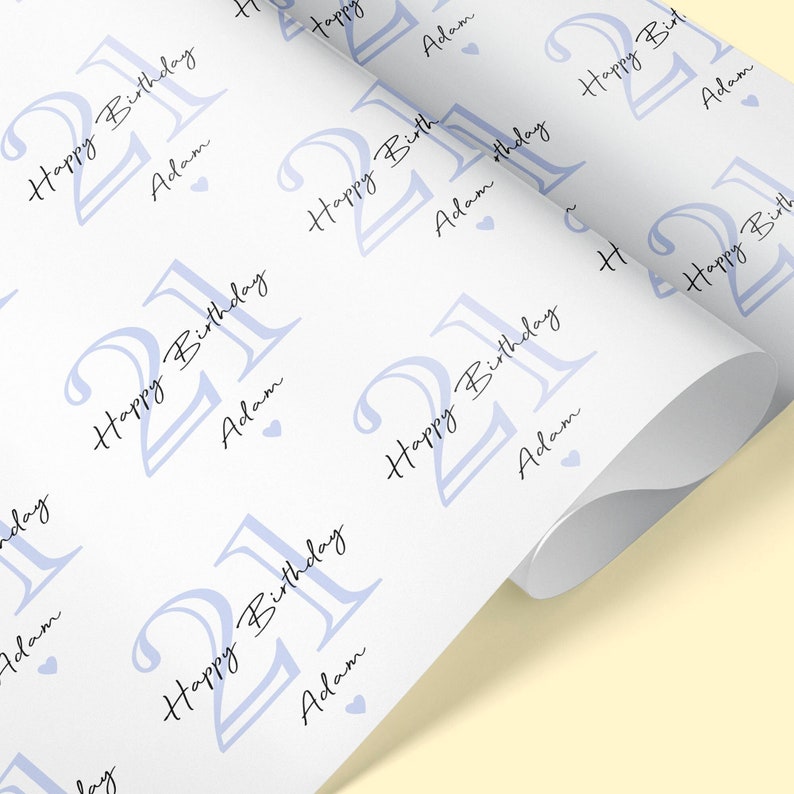 May include: White wrapping paper with a repeating pattern of the number "21" in light blue, the words "Happy Birthday" and the name "Adam" in black script. Small blue hearts are scattered throughout the design. The paper is rolled.
