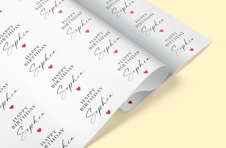 Pode incluir: Papel de embrulho branco impresso com a frase "HAPPY BIRTHDAY Sophia" em uma fonte elegante, acompanhada de pequenos &iacute;cones de cora&ccedil;&atilde;o vermelho. O papel est&aacute; parcialmente enrolado, revelando o padr&atilde;o.