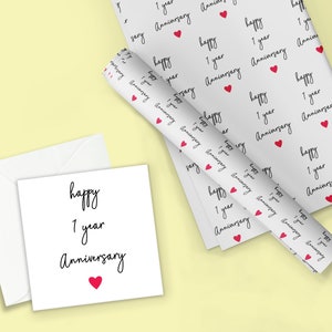 Op de afbeelding: Een witte wenskaart en envelopset met de woorden "happy 1 year Anniversary" in zwart schrift en een rood hart. Een rol bijpassend cadeaupapier en een plat vel zijn ook zichtbaar, met dezelfde tekst en hartpatroon.