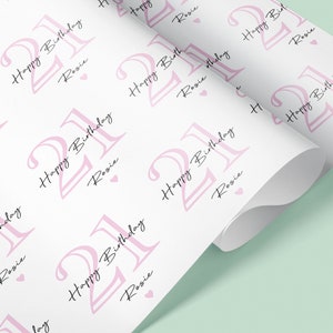 May include: White wrapping paper with a repeating pattern of the number "21" in pink, the words "Happy Birthday" and the name "Rosie" in a script font, and small pink hearts. The paper is rolled up.