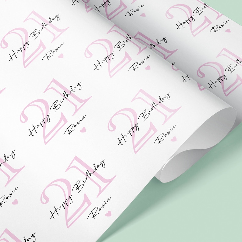 May include: White wrapping paper with a repeating pattern of the number "21" in pink, the words "Happy Birthday" and the name "Rosie" in a script font, and small pink hearts. The paper is rolled up.
