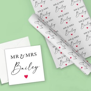 Op de afbeelding: Wit cadeaupapier, een kaart en een envelop met de tekst "MR & MRS Bailey" in zwart script, geaccentueerd met rode hartjes. Het cadeaupapier is opgerold en plat, met hetzelfde patroon. De kaart is vierkant.
