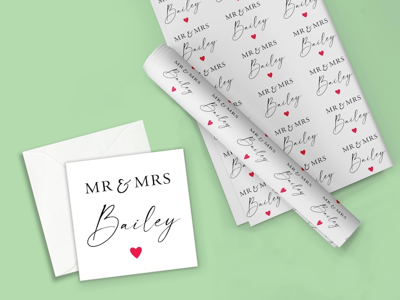 Op de afbeelding: Wit cadeaupapier, een kaart en een envelop met de tekst "MR & MRS Bailey" in zwart script, geaccentueerd met rode hartjes. Het cadeaupapier is opgerold en plat, met hetzelfde patroon. De kaart is vierkant.