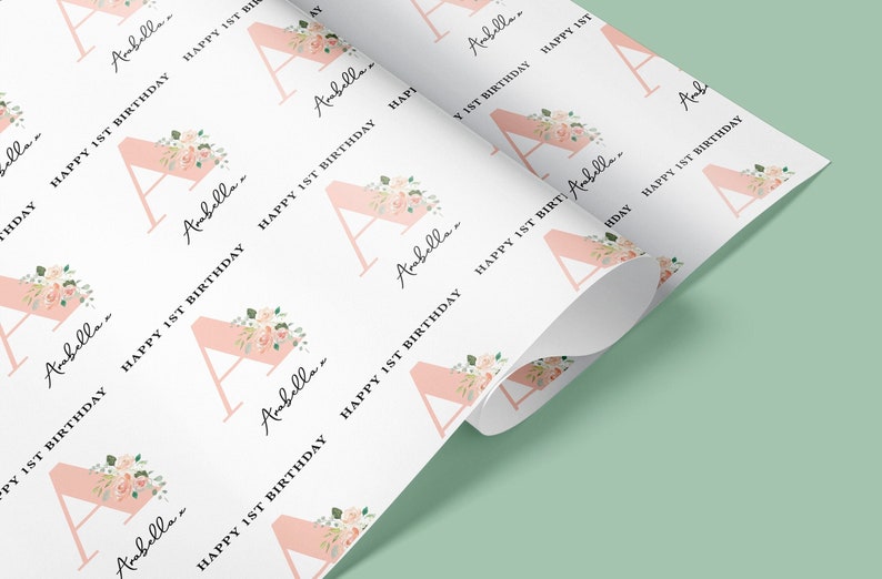 May include: White wrapping paper with a repeating pattern of a pink letter A with floral accents and the text "Arabella... Happy 1st Birthday".