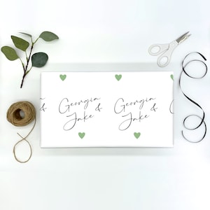 Pu&ograve; includere: Carta da regalo bianca con un motivo ripetuto di cuori verde chiaro e i nomi "Georgia & Jake" in corsivo nero. Una bobina di spago, forbici, un rametto di verde e nastro nero sono disposti attorno al regalo confezionato.