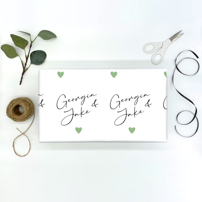 Pu&ograve; includere: Carta da regalo bianca con un motivo ripetuto di cuori verde chiaro e i nomi "Georgia & Jake" in corsivo nero. Una bobina di spago, forbici, un rametto di verde e nastro nero sono disposti attorno al regalo confezionato.