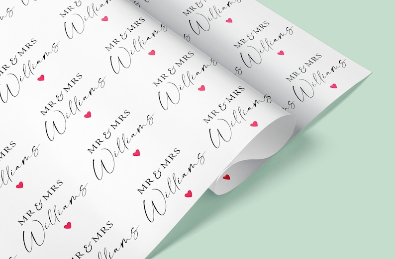 Op de afbeelding: Wit cadeaupapier met een herhalend patroon van "MR & MRS Williams" in een handschrift, geaccentueerd met kleine rode hartjes. Het papier is opgerold en toont het ontwerp aan de bovenkant en binnenkant.