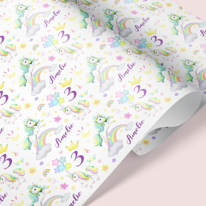 Op de afbeelding: Wit inpakpapier met een herhalend patroon van kleurrijke eenhoorns, regenbogen, sterren en bloemen. De naam "Amelie" en het nummer "3" worden herhaald in het hele patroon.