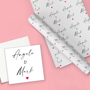 K&ouml;nnte beinhalten: Wei&szlig;es Geschenkpapier mit einem sich wiederholenden Muster der Namen "Angela & Mark" in schwarzer Schrift mit einem kleinen roten Herzen zwischen jedem Namen.