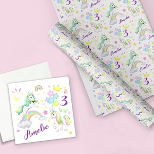 Op de afbeelding: Een witte kaart met een ontwerp van een eenhoorn en regenboog. De kaart heeft de naam "Amelie" en het nummer "3" erop. De kaart wordt vergezeld door een witte envelop.