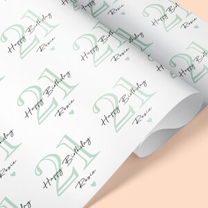 May include: White wrapping paper with a repeating pattern of the number 21 in a light green color, along with the words "Happy Birthday Rosie" in black script. Small green hearts are scattered throughout the design.