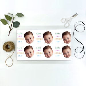 May include: White wrapping paper with nine colorful stickers featuring a baby's face and the text "Happy First Birthday Eden".