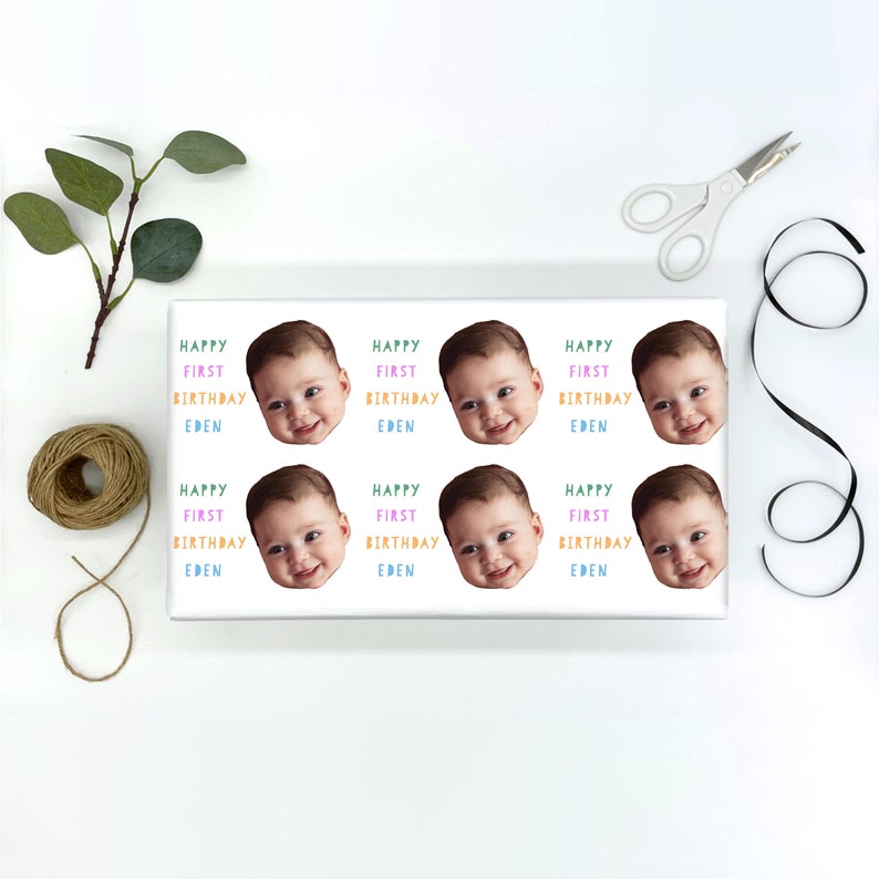 May include: White wrapping paper with nine colorful stickers featuring a baby's face and the text "Happy First Birthday Eden".