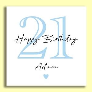 May include: A white birthday card with a light blue "21" and the words "Happy Birthday Adam" in black script. A small blue heart is at the bottom. The card is on a yellow background.