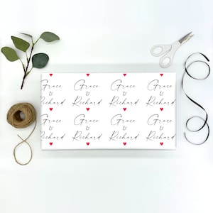 K&ouml;nnte beinhalten: Wei&szlig;es Geschenkpapier mit einem sich wiederholenden Muster aus roten Herzen und dem Text "Grace & Richard" in schwarzer Schrift.