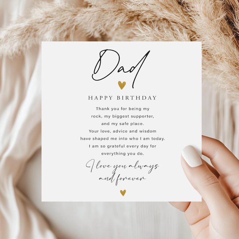 May include: A white card with the word "Dad" in elegant script, accompanied by "Happy Birthday" and a heartfelt message. Gold heart accents add a touch of warmth. The card is held by a person.