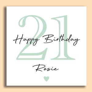 May include: A white birthday card with the number 21 in a soft green color. The words "Happy Birthday" and the name "Rosie" are written in black script. A small green heart is at the bottom.