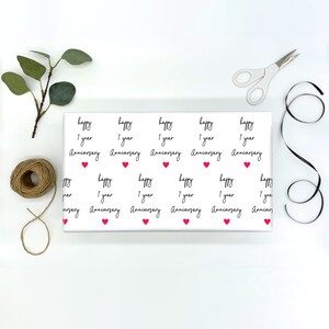 Op de afbeelding: Wit cadeaupapier met een herhaald patroon van de woorden "happy 1 year Anniversary" in zwart schrift, afgewisseld met kleine rode hartjes. Een witte schaar, zwart lint en touw zijn rond het papier gerangschikt.