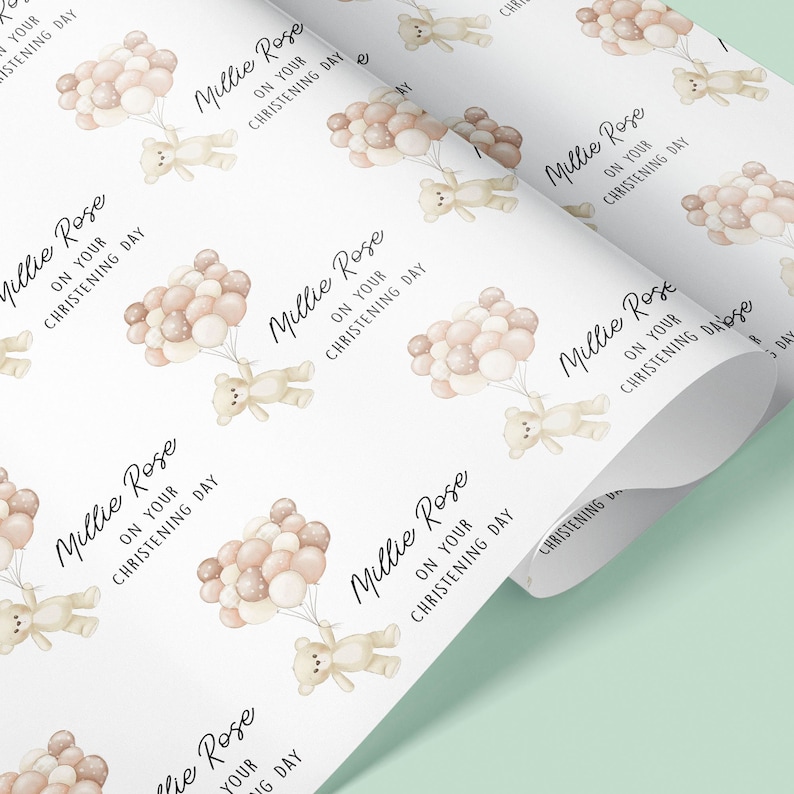 K&ouml;nnte beinhalten: Wei&szlig;es Geschenkpapier mit einem sich wiederholenden Muster aus einem Teddyb&auml;r, der Ballons h&auml;lt, auf denen "Millie Rose on your christening day" steht.