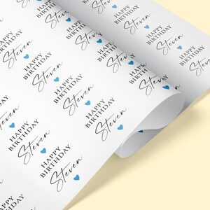 Pode incluir: Papel de embrulho branco impresso com o texto "HAPPY BIRTHDAY Steven" em uma fonte elegante, repetido na superf&iacute;cie. Pequenos &iacute;cones de cora&ccedil;&atilde;o azul s&atilde;o intercalados no design. O papel est&aacute; enrolado.