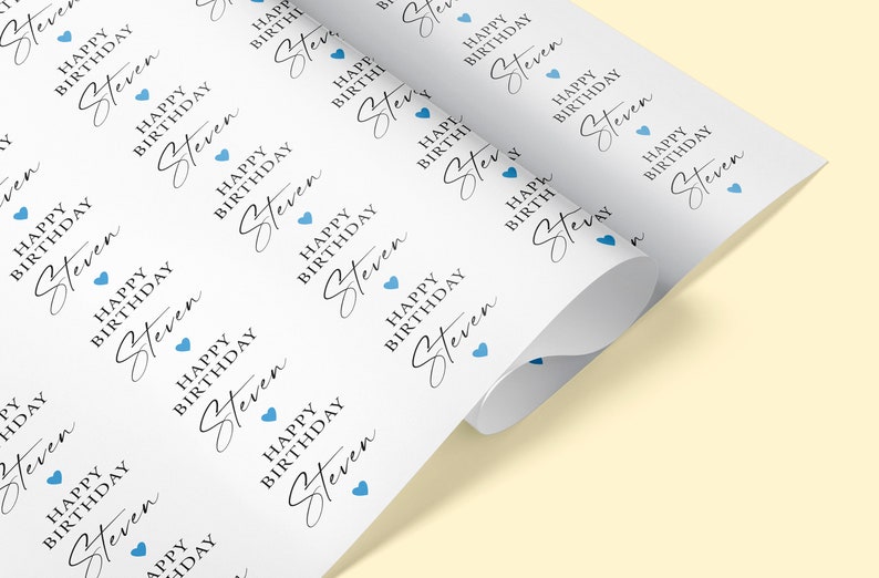 Pode incluir: Papel de embrulho branco impresso com o texto "HAPPY BIRTHDAY Steven" em uma fonte elegante, repetido na superf&iacute;cie. Pequenos &iacute;cones de cora&ccedil;&atilde;o azul s&atilde;o intercalados no design. O papel est&aacute; enrolado.