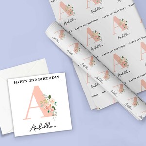 May include: A white card with pink floral design and the text "Happy 2nd Birthday" and "Arabella" with a pink letter A. A roll of white wrapping paper with a pink floral design and the text "Happy 1st Birthday" and "Arabella" with a pink letter A.