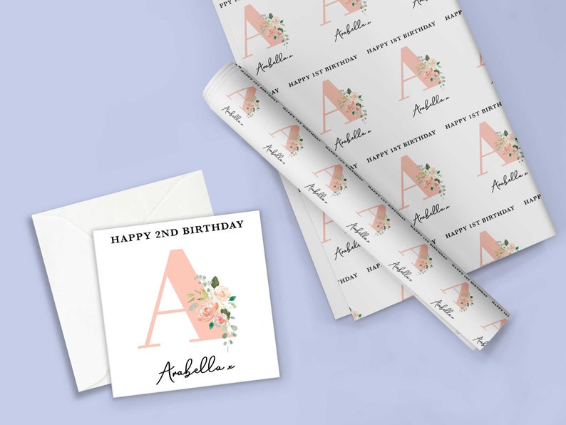 May include: A white card with pink floral design and the text "Happy 2nd Birthday" and "Arabella" with a pink letter A. A roll of white wrapping paper with a pink floral design and the text "Happy 1st Birthday" and "Arabella" with a pink letter A.
