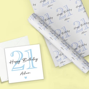 May include: A birthday gift set featuring a white card with a light blue "21 Happy Birthday Adam" design, a matching envelope, and wrapping paper with a repeating pattern of the same design. The wrapping paper is rolled up.