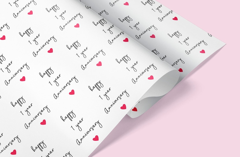 Op de afbeelding: Wit cadeaupapier met een patroon van de woorden "happy 1 year Anniversary" in zwart schrift, afgewisseld met kleine rode hartjes. Het papier is gedeeltelijk opgerold.