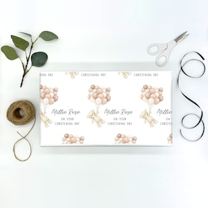 K&ouml;nnte beinhalten: Wei&szlig;es Geschenkpapier mit einem sich wiederholenden Muster aus einem Teddyb&auml;r, der Ballons h&auml;lt. Der Text "Millie Rose on your christening day" ist auf dem Papier gedruckt.