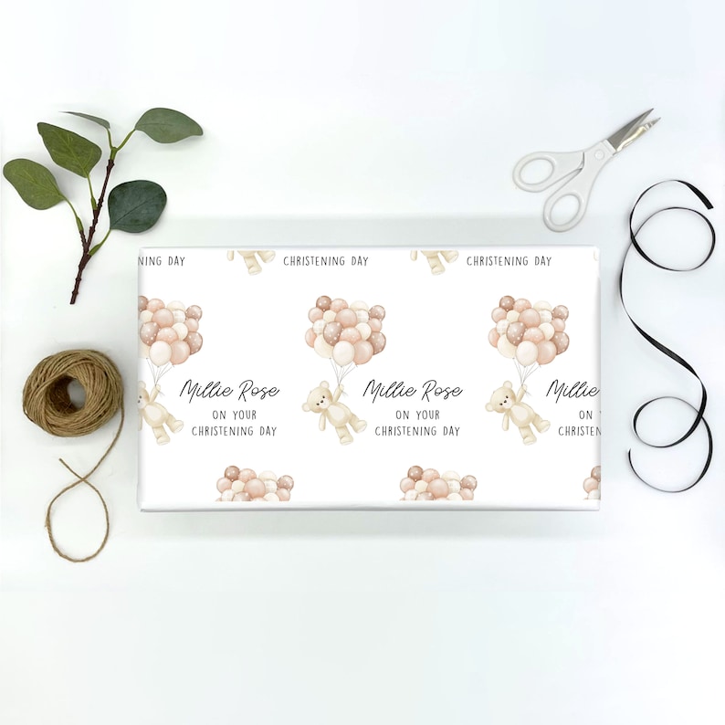 K&ouml;nnte beinhalten: Wei&szlig;es Geschenkpapier mit einem sich wiederholenden Muster aus einem Teddyb&auml;r, der Ballons h&auml;lt. Der Text "Millie Rose on your christening day" ist auf dem Papier gedruckt.